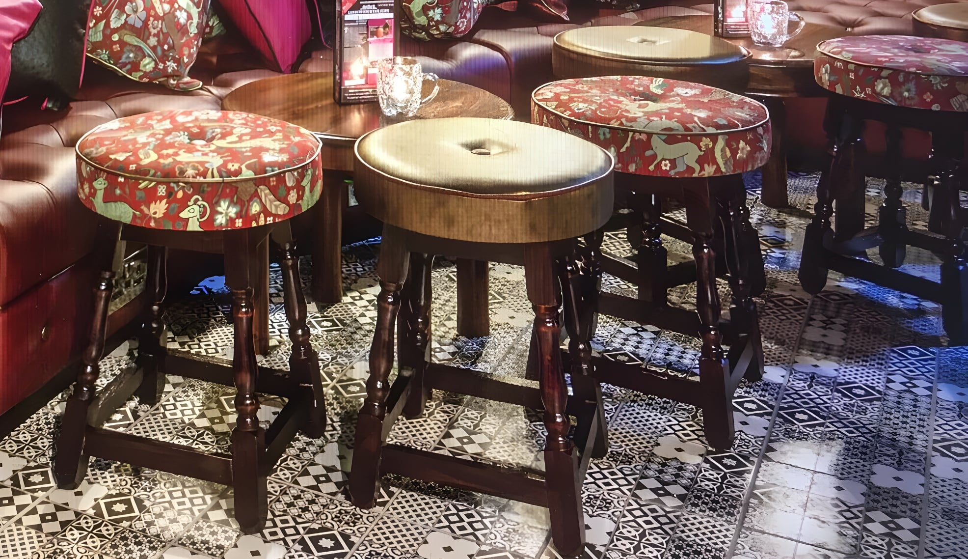 Turned dark wood stools re-covered in a red floral tapestry and gold vinyl, set on patterned tiles in a bar.
