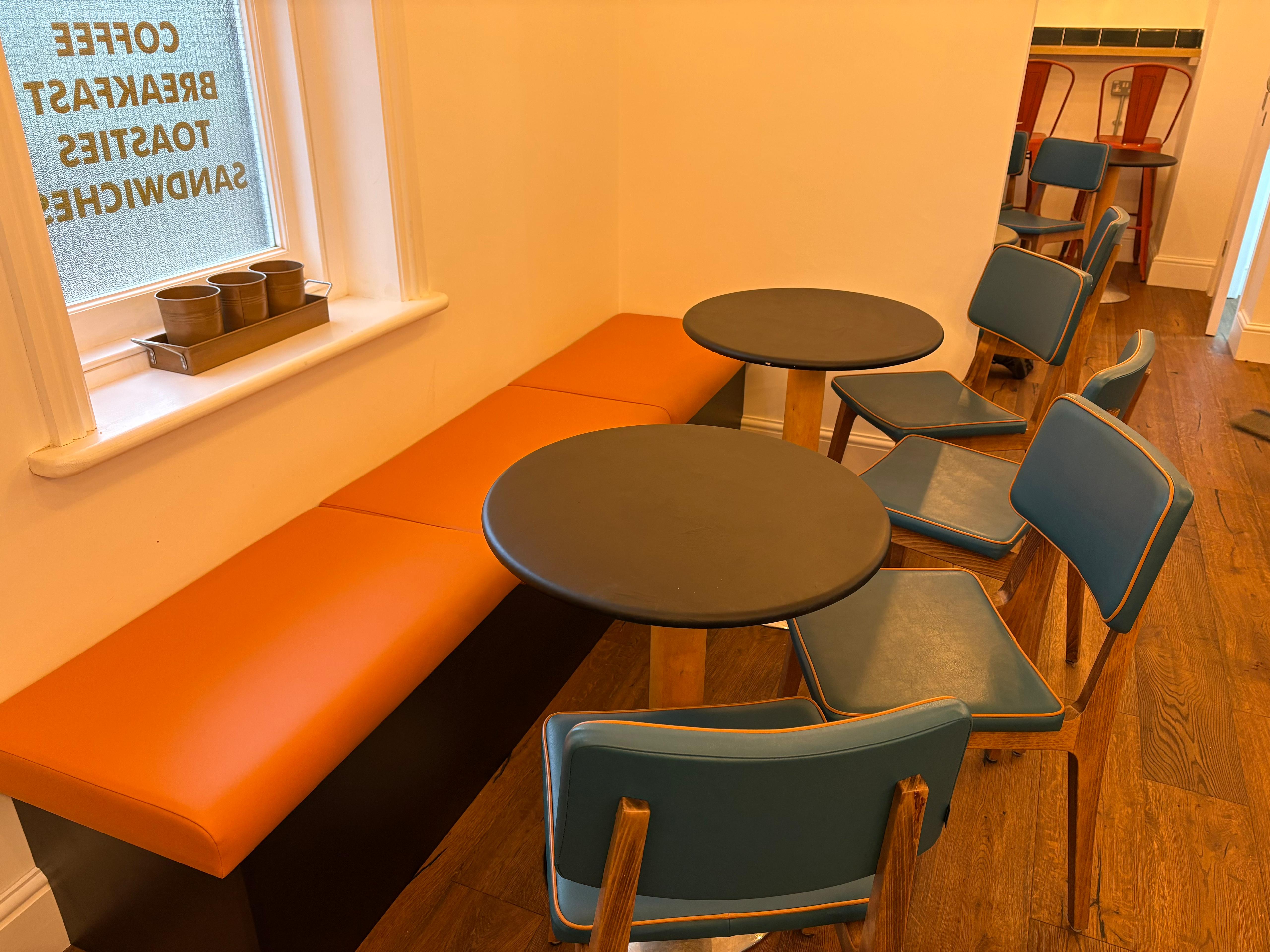 Slim orange faux leather bench running under a cafe window, paired with teal piped chairs and round tables.
