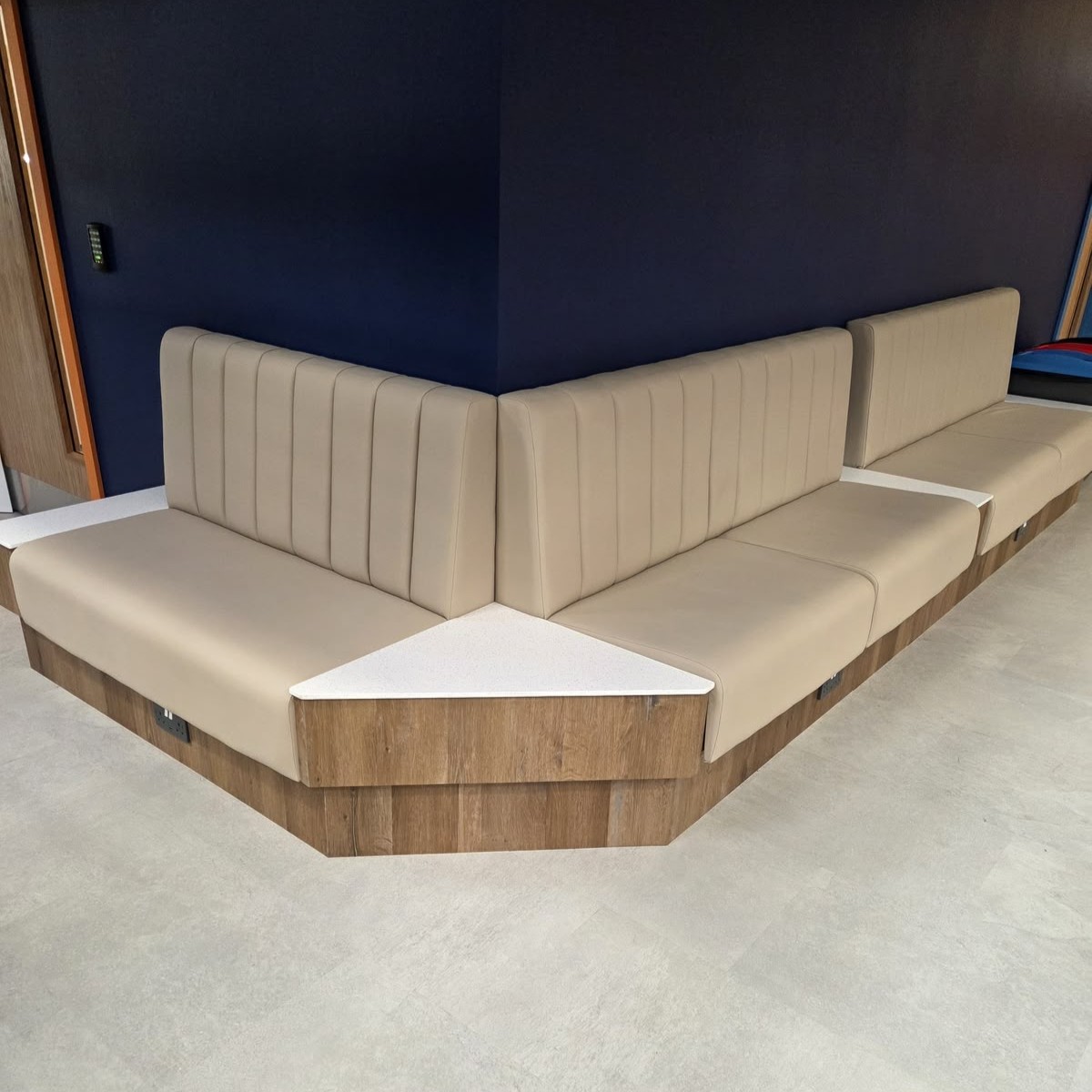 L-shaped fluted booth in beige leather on an oak plinth with an inset stone corner table, built for a sports venue lounge.