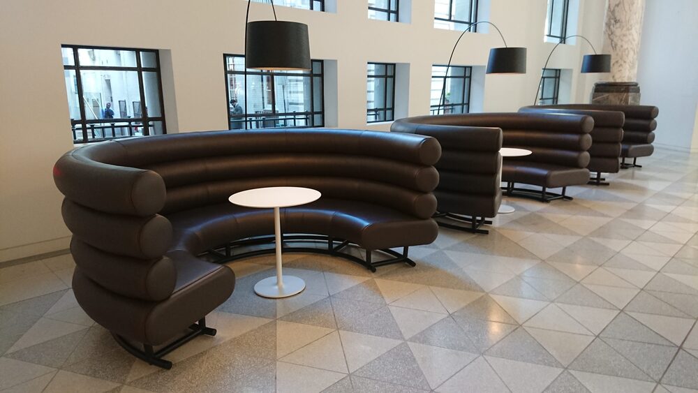 A run of semi-circular sofas in brown leather with stacked tubular backs, set on steel sled bases in a corporate atrium.