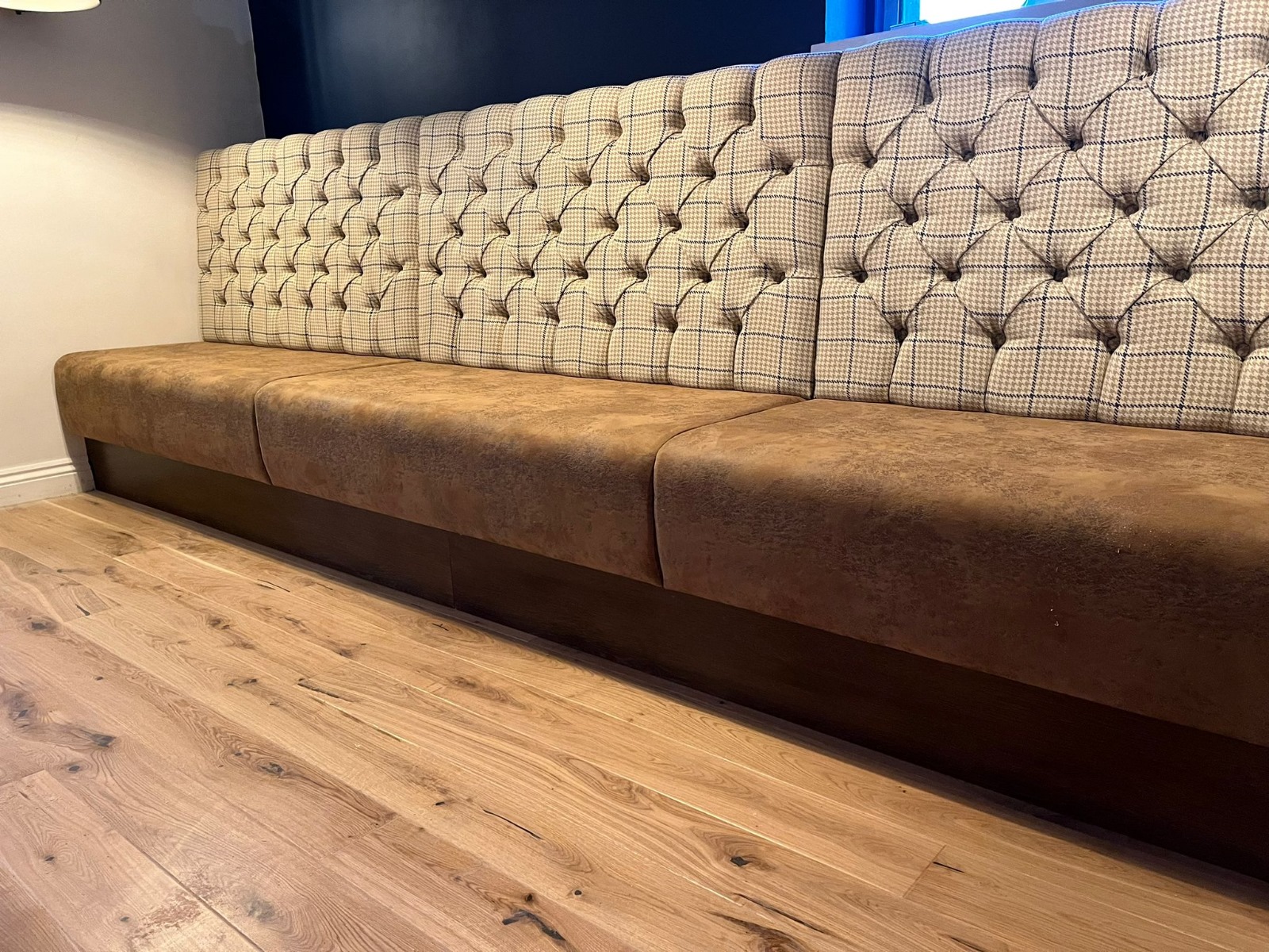 Deep-buttoned check fabric backrest above a distressed brown suede seat, set on a dark timber plinth.