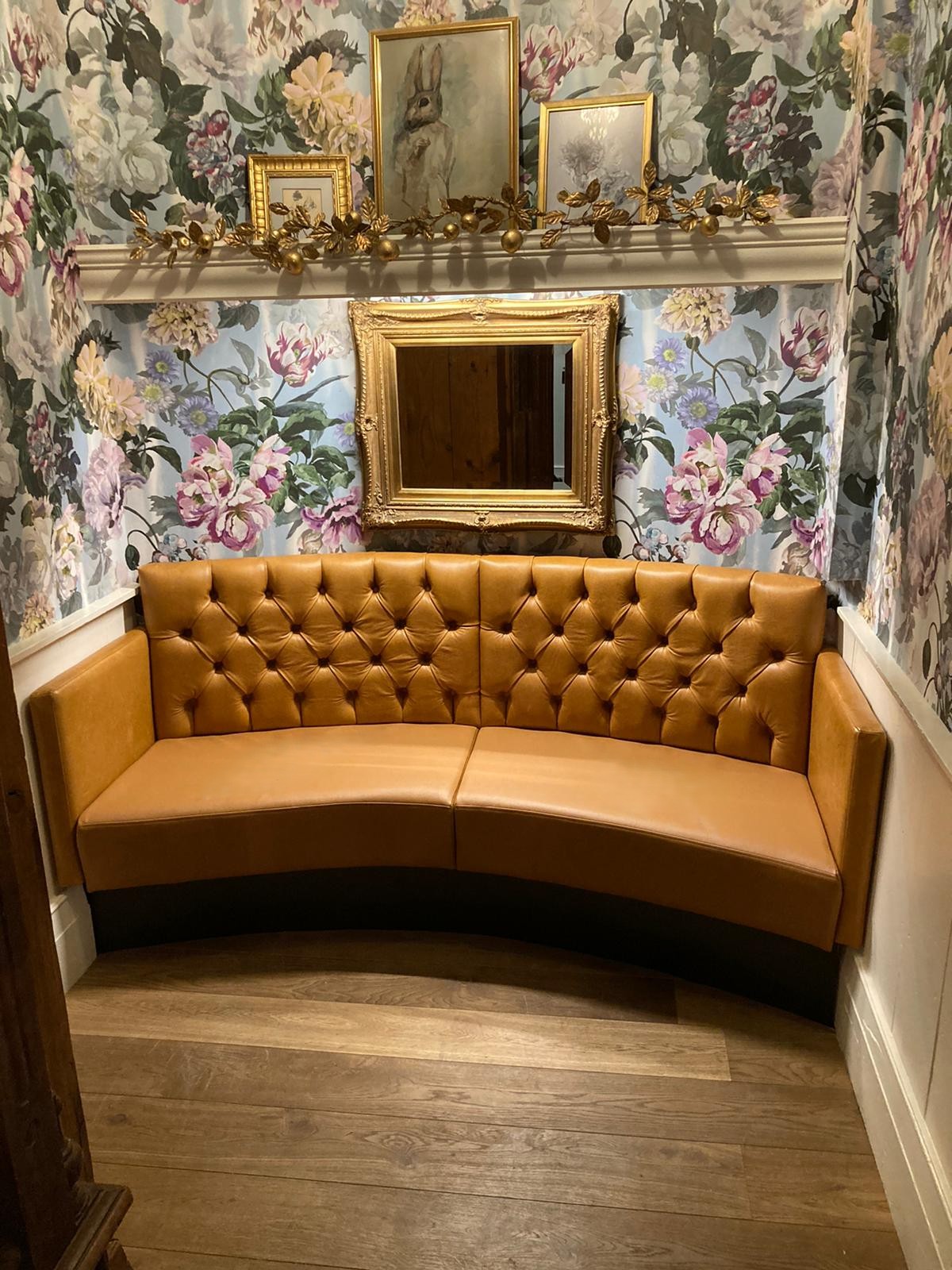 Curved deep-buttoned banquette in tan leather set into a floral-papered alcove beneath a gilt mirror.