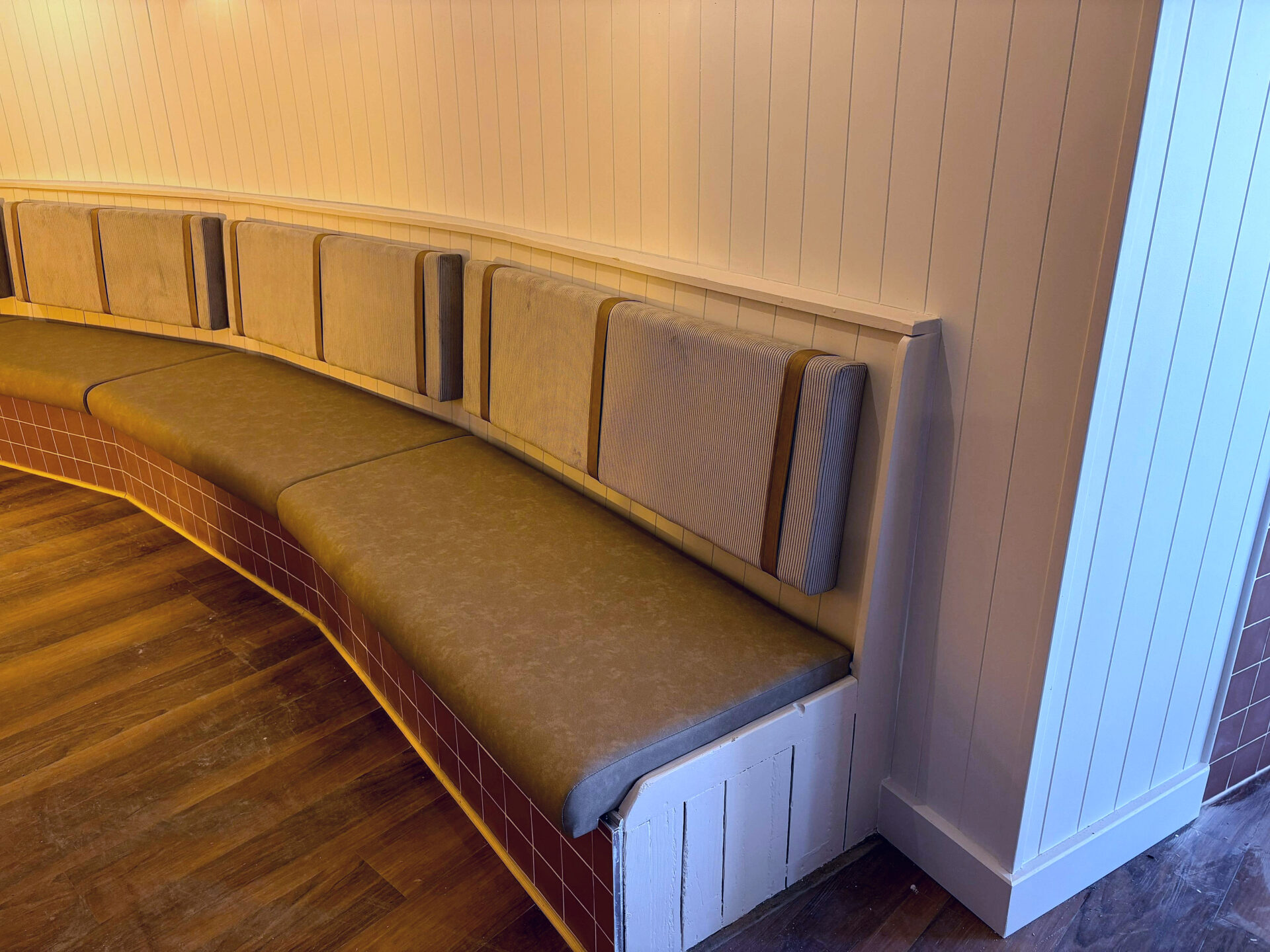 Curved wall banquette with a tan suede seat and individual corduroy-trimmed back panels, following a tiled plinth around the dining room.