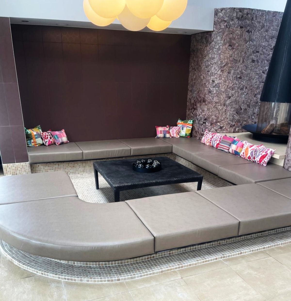 Sunken lounge with taupe leather modular cushions built into a mosaic-tiled plinth around a central fire.