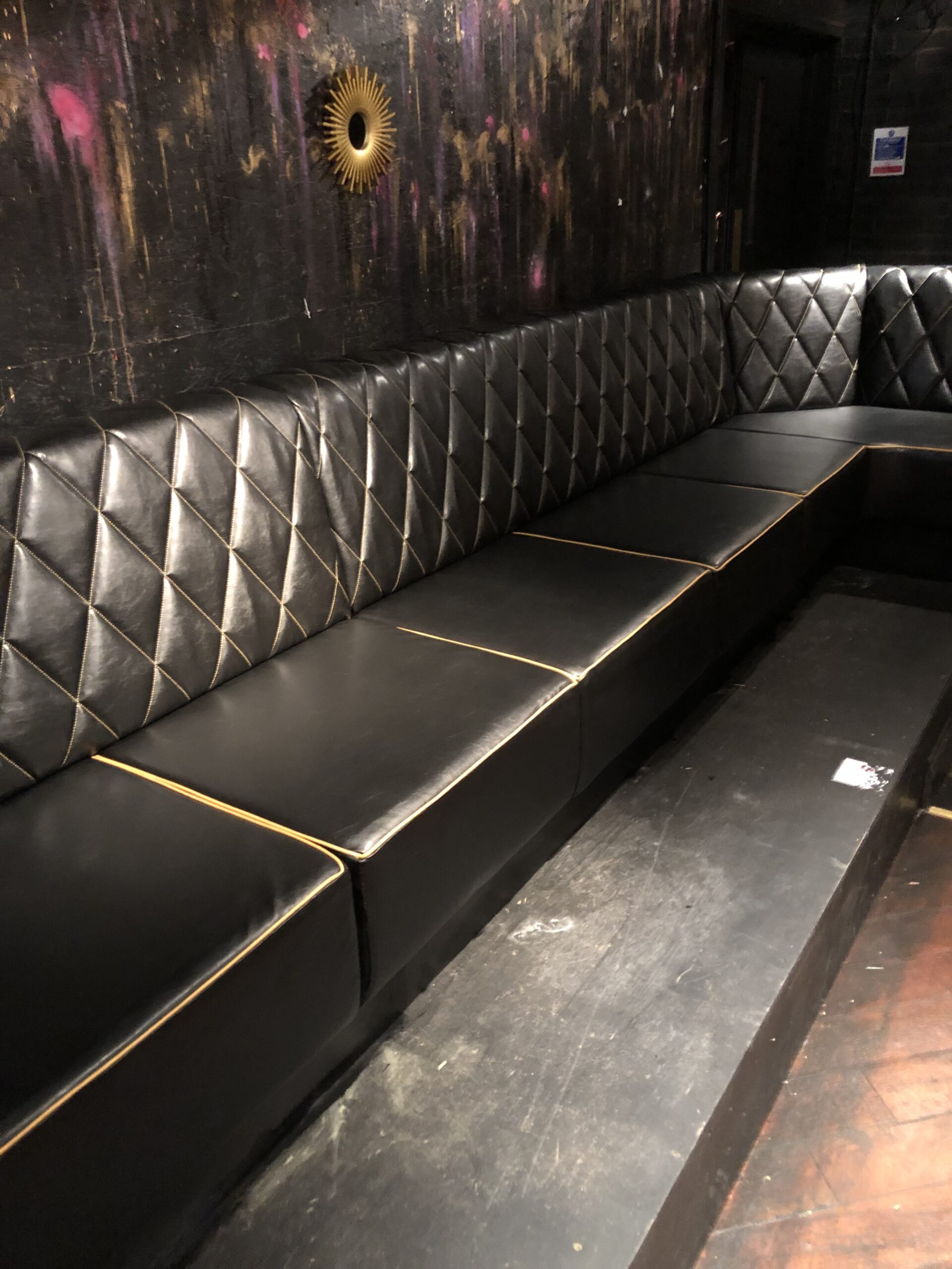 Wrap-around black leather banquette with gold diamond stitching and contrast piping, fitted along the back wall of a club.
