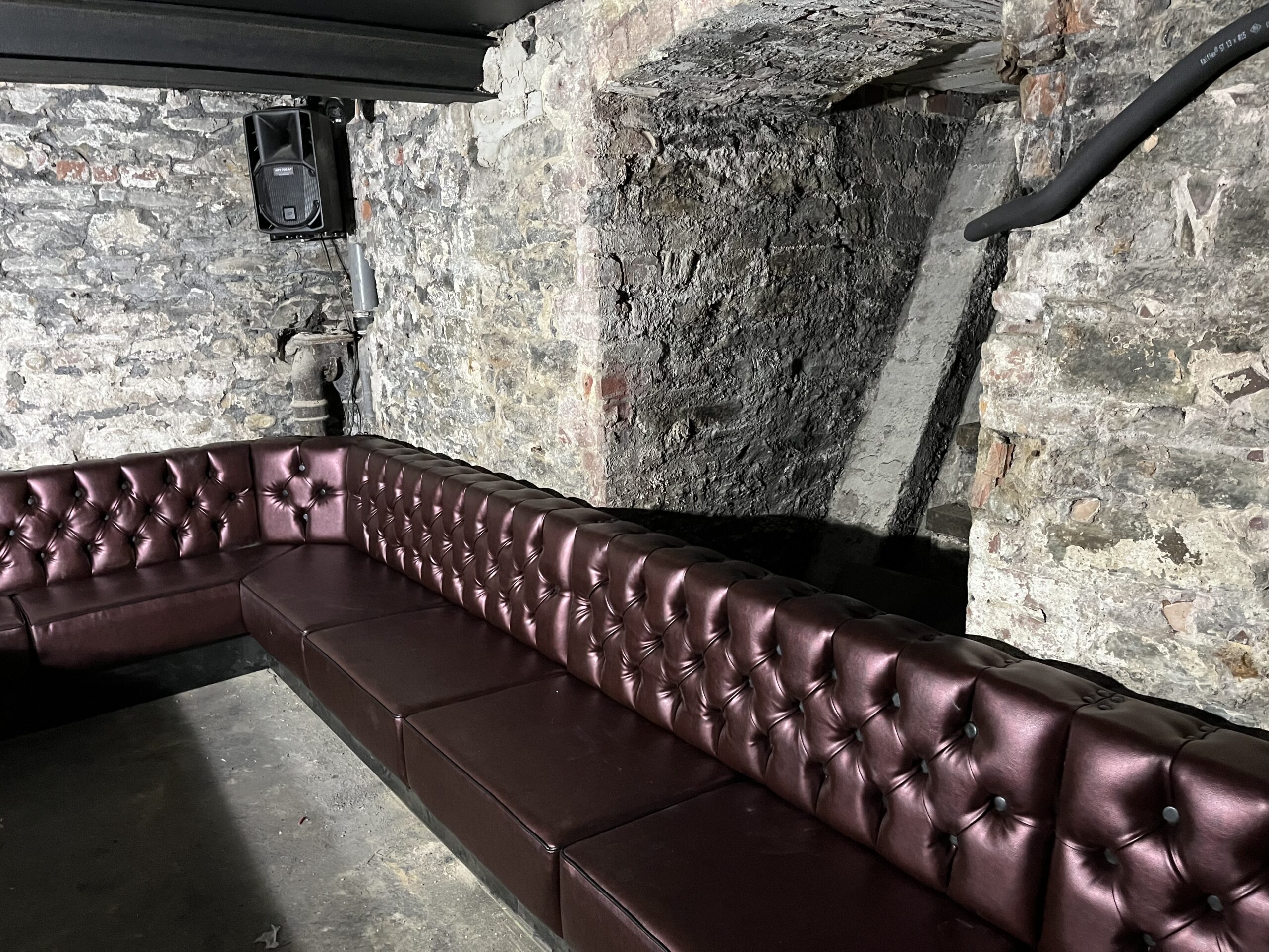 Deep-buttoned burgundy leatherette banquette running along the exposed stone walls of a cellar club.