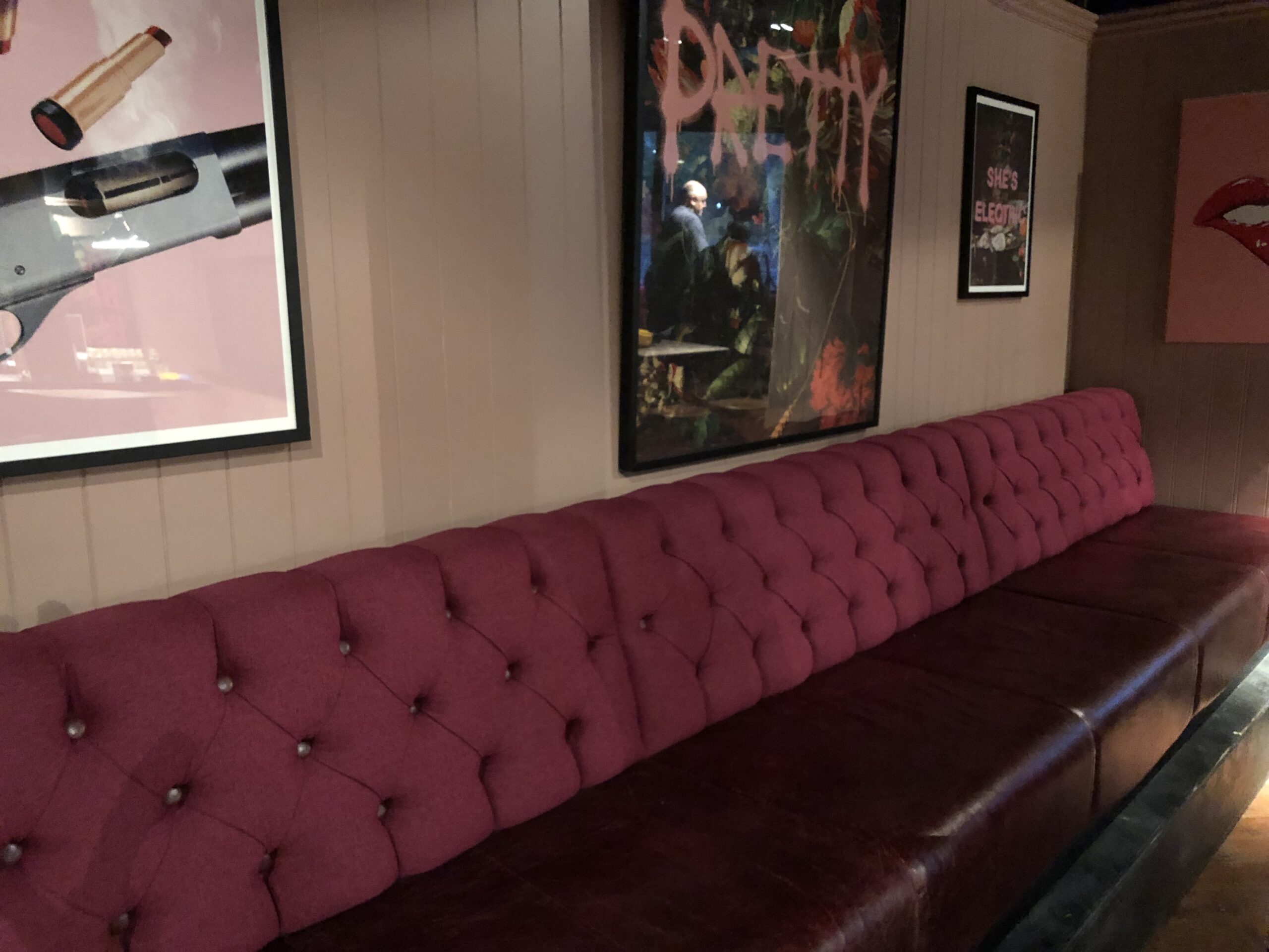 Long deep-buttoned banquette in raspberry wool over a burgundy leather seat, running beneath framed artwork in a bar.