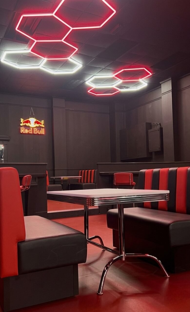 Back-to-back diner booths in red and black vinyl with chrome-edged tables, under hexagon neon lighting.