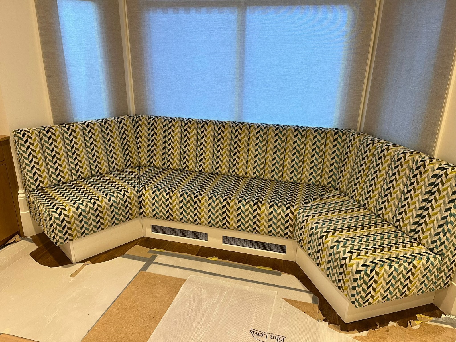 Bay-window corner bench upholstered in a green and ochre chevron weave, with fluted backs and a painted plinth.