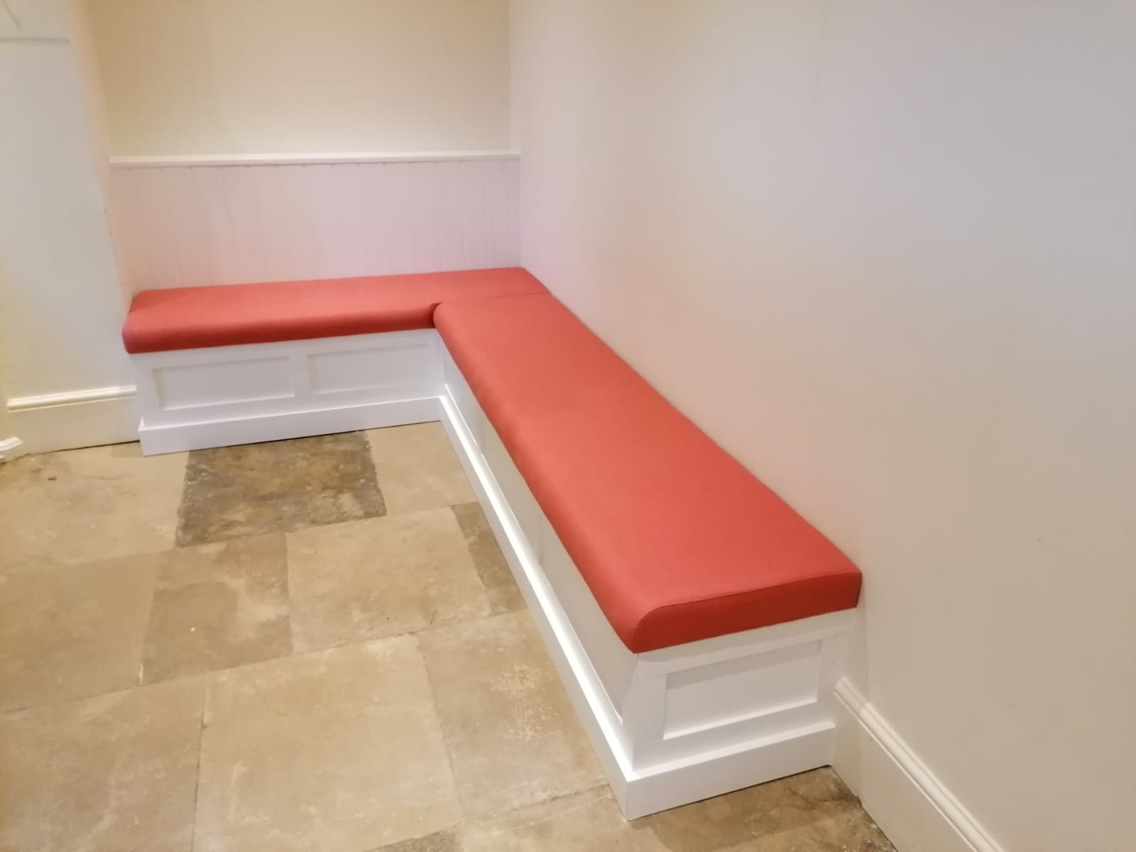 L-shaped bench with a coral wool seat pad set into painted panelled joinery, built into a flagstone hallway corner.
