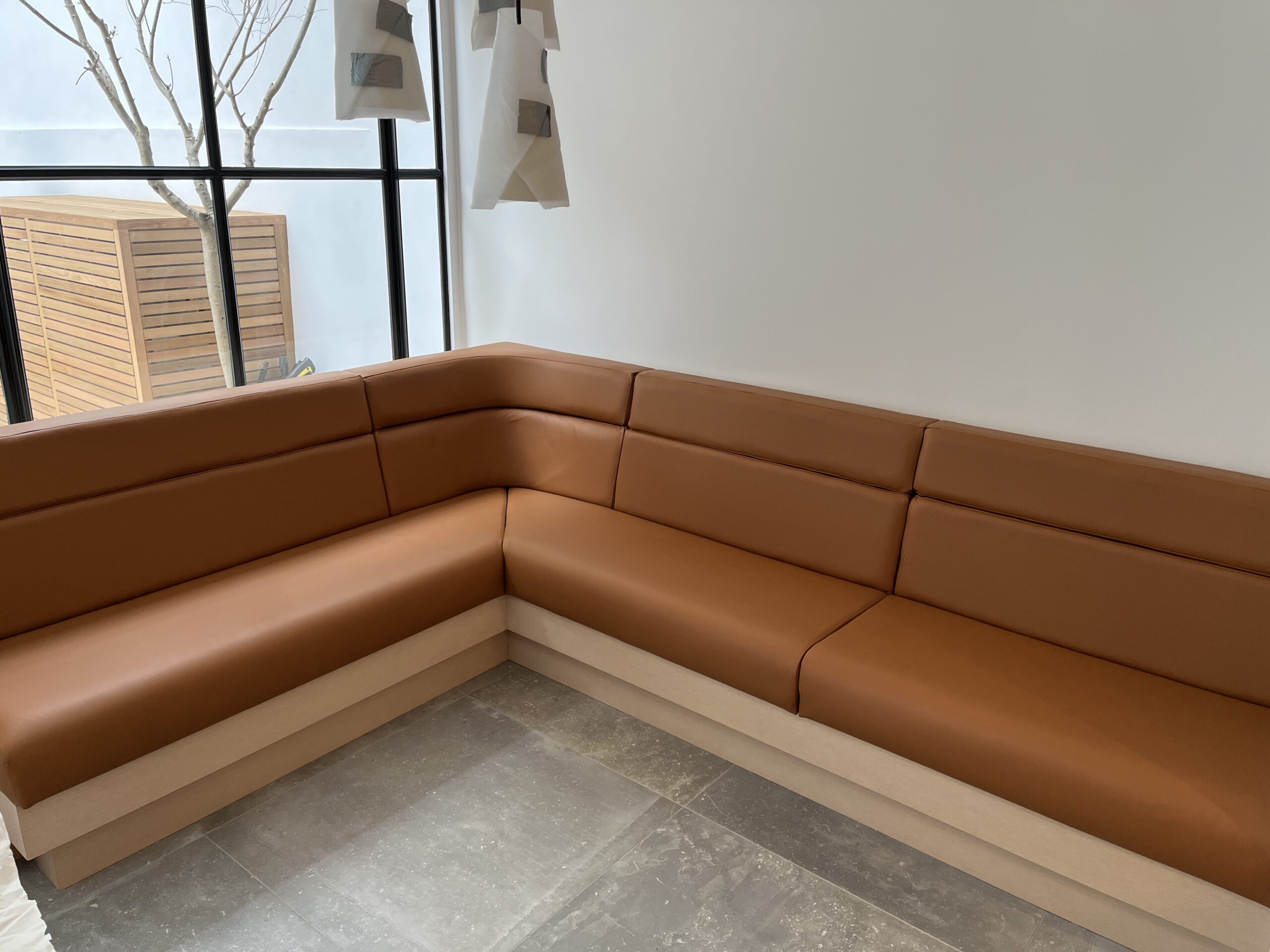 Minimal corner banquette in warm tan leather on a pale oak plinth, wrapping a full-height window.