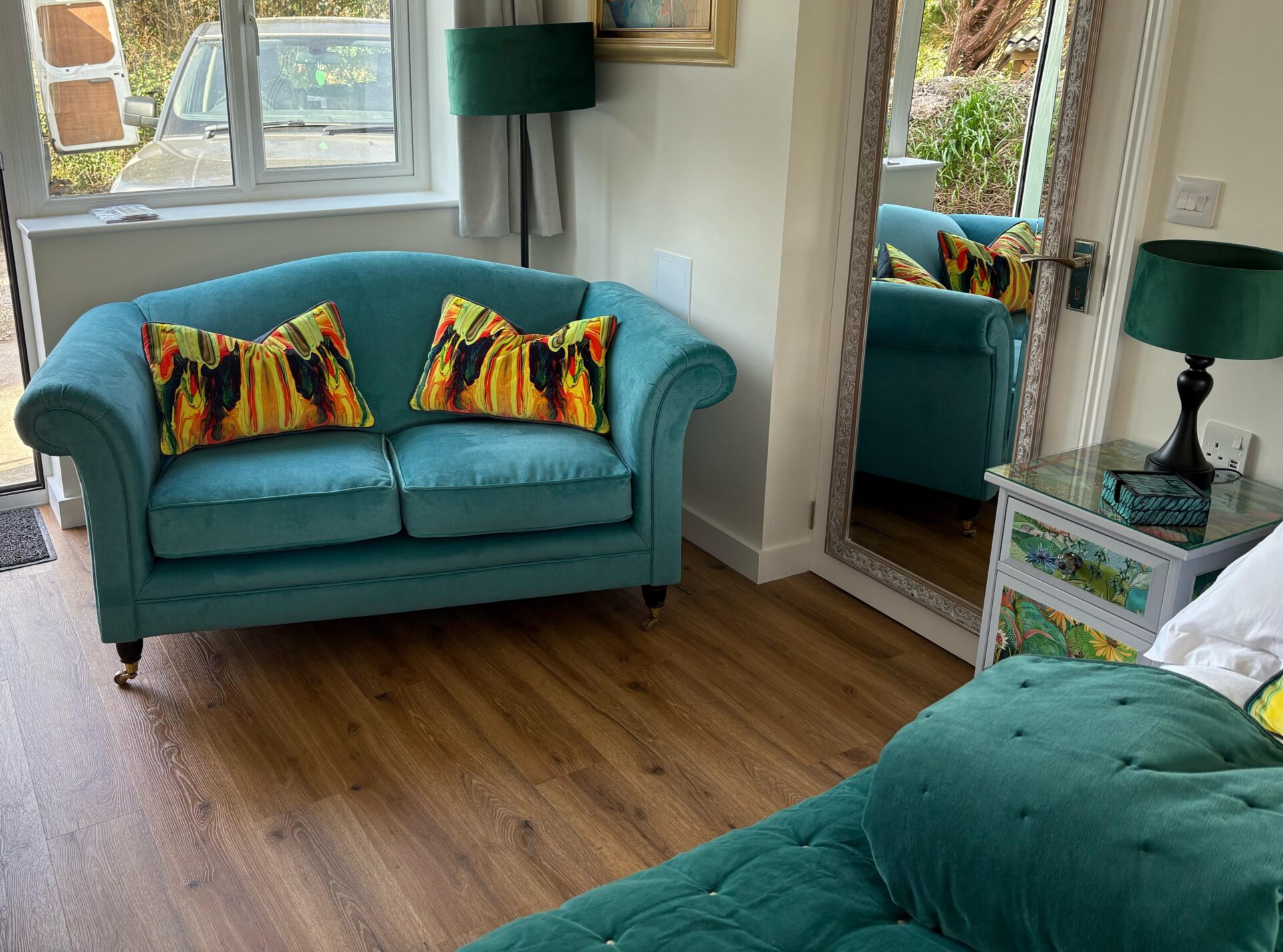 Two-seater velvet sofa in teal with rolled arms and turned legs on brass castors, finished with bold hand-painted scatter cushions.