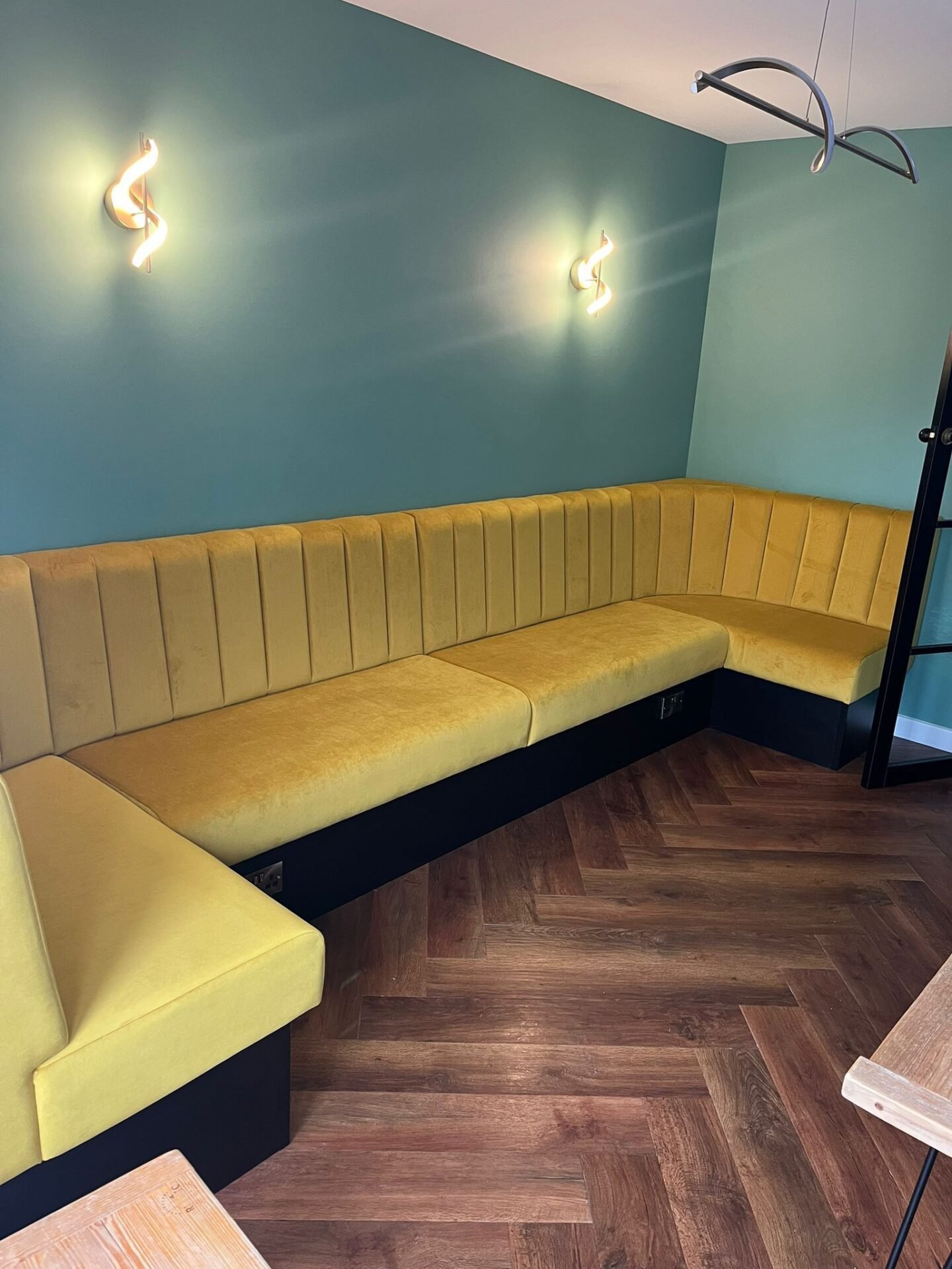 U-shaped banquette in ochre velvet with a fluted back on a black plinth, set against teal walls and herringbone flooring.