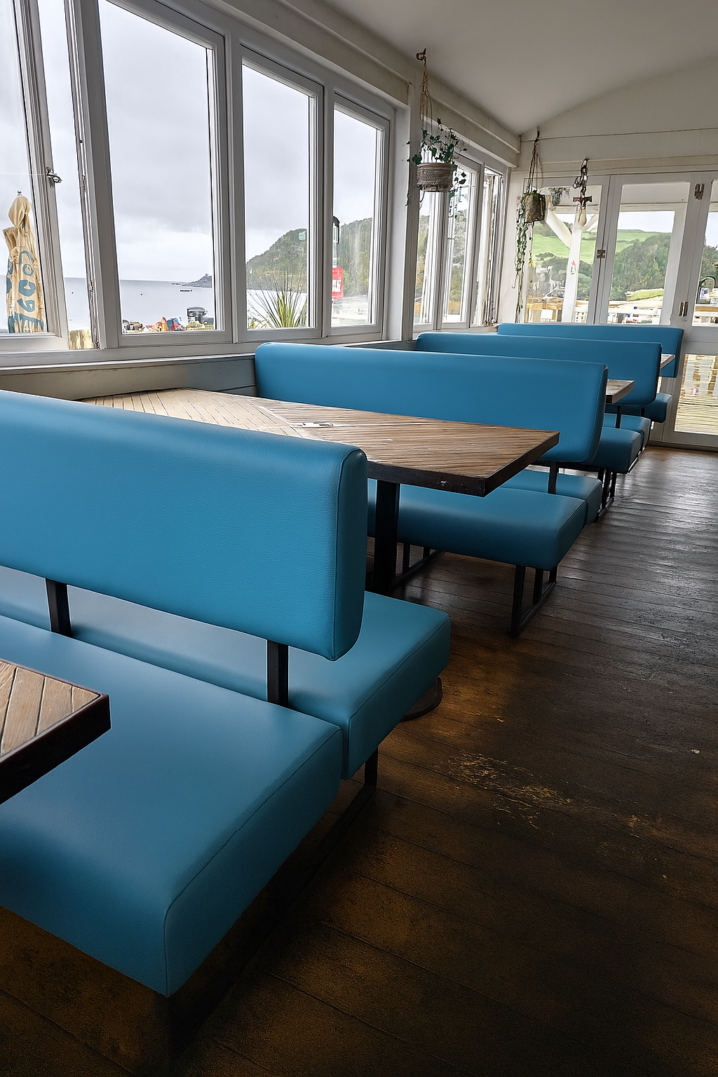 Rows of low blue faux leather booths on black steel frames, set against the windows of a coastal cafe.