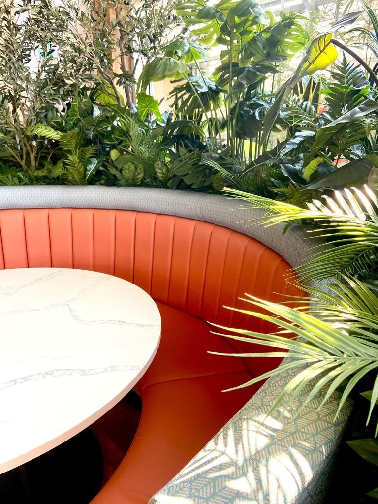 Curved fluted banquette in burnt orange leather topped with a woven grey capping, wrapped by planting in a garden centre restaurant.