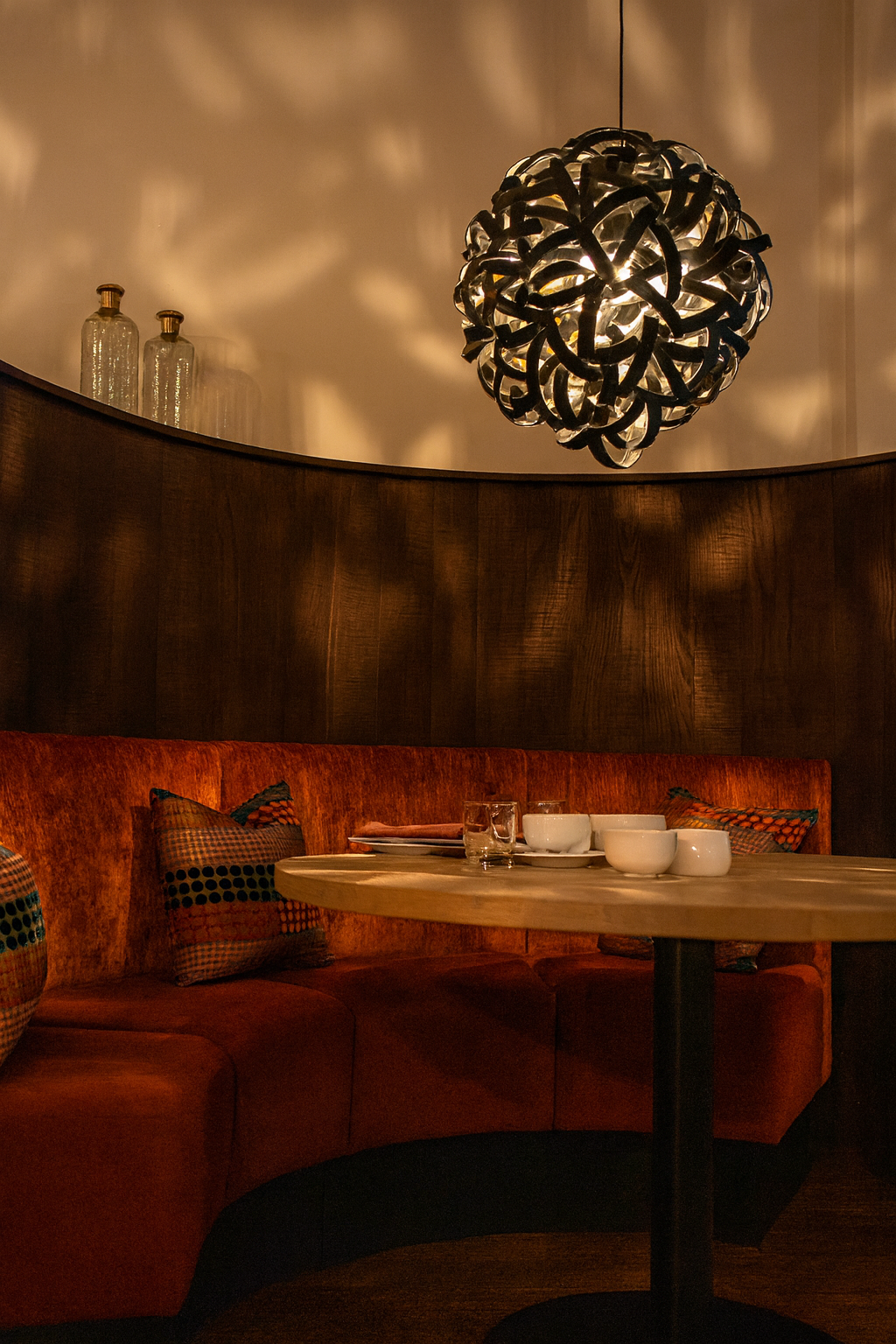 Curved banquette in burnt orange velvet and suede against charred timber panelling, dressed with retro spot cushions.