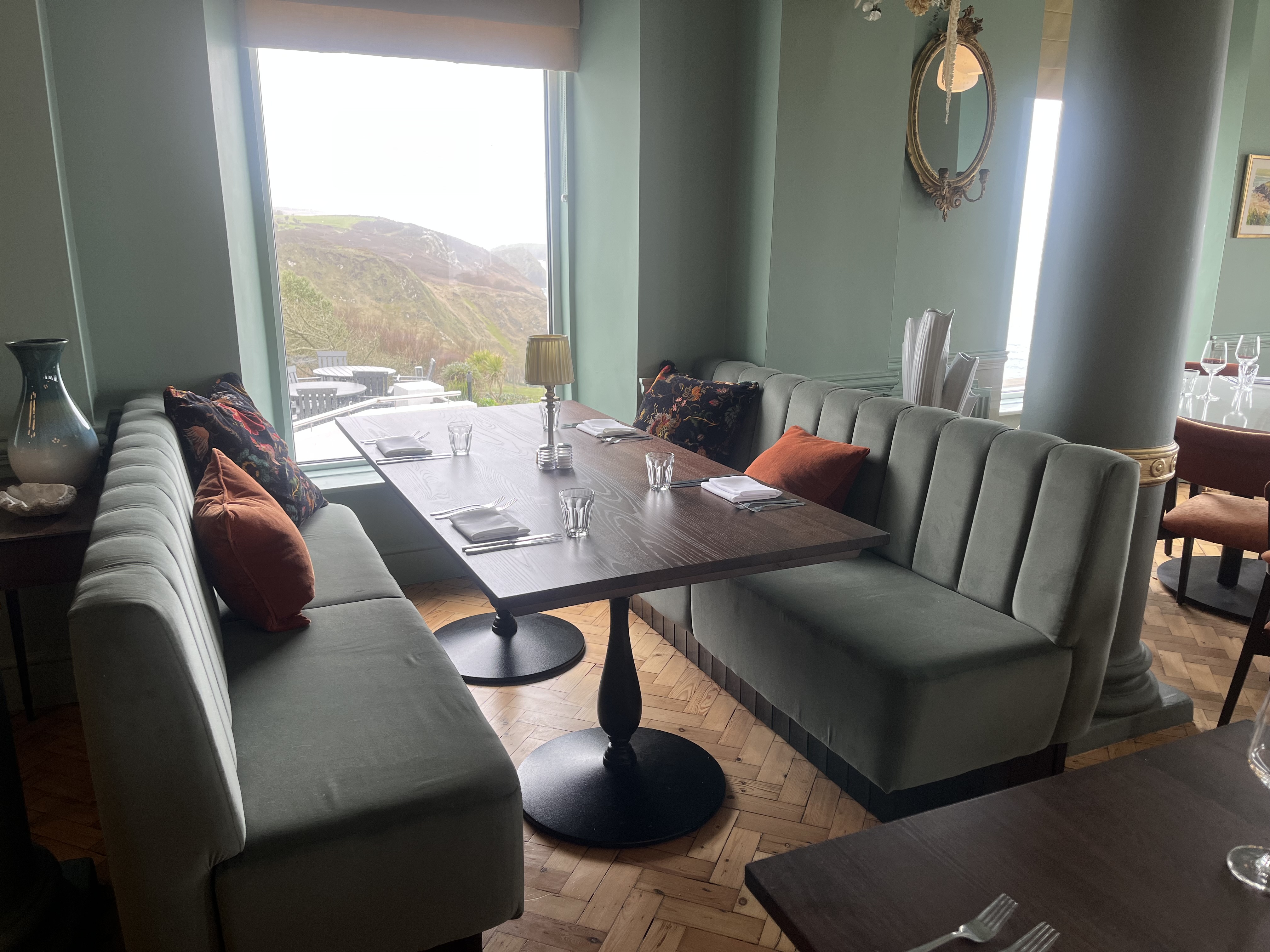 Facing fluted banquettes in sage green velvet set either side of a dark oak table, looking out over the Cornish coast.