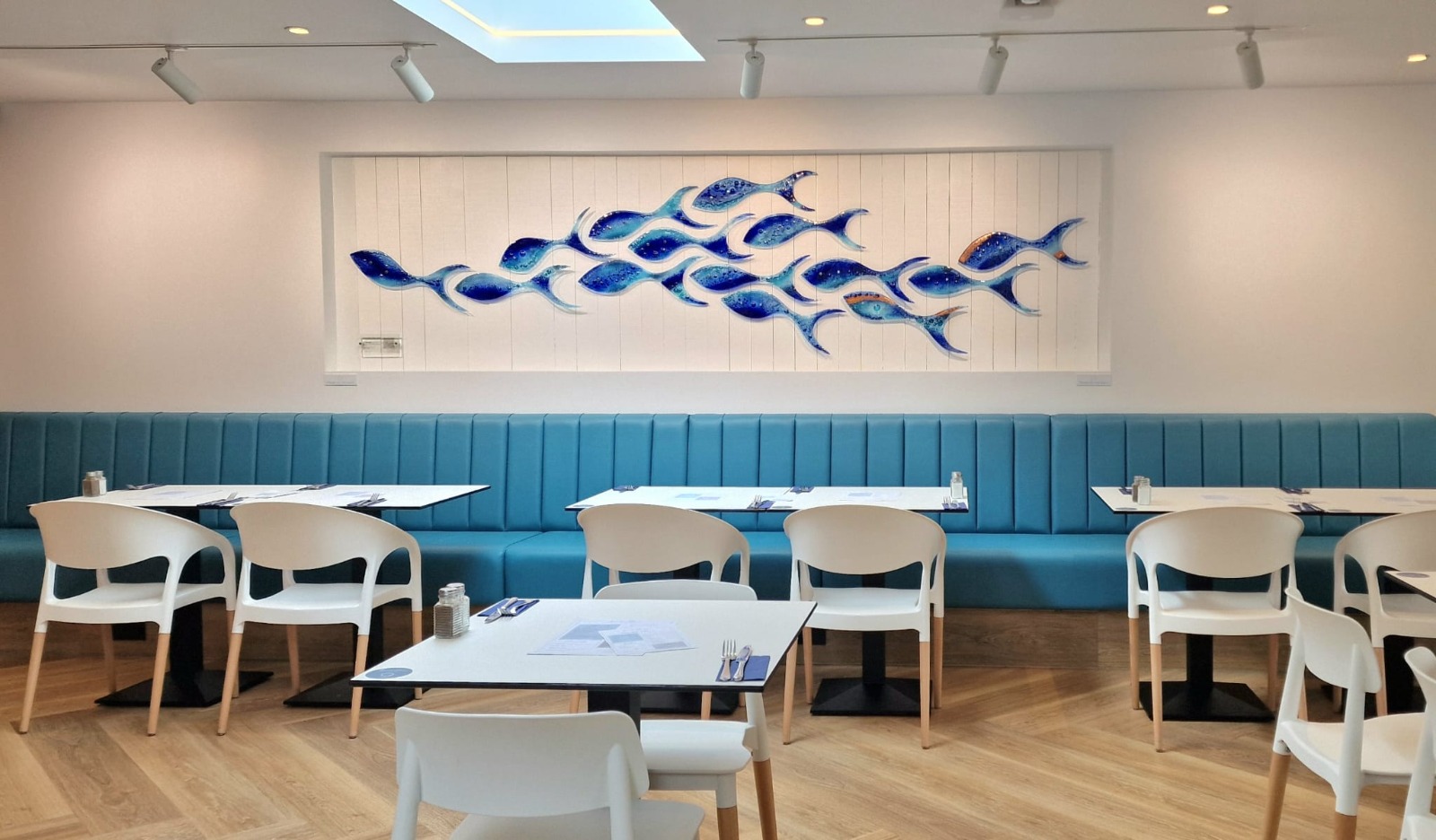 Long teal fluted banquette running the width of a bright dining room beneath a shoal of glass fish.